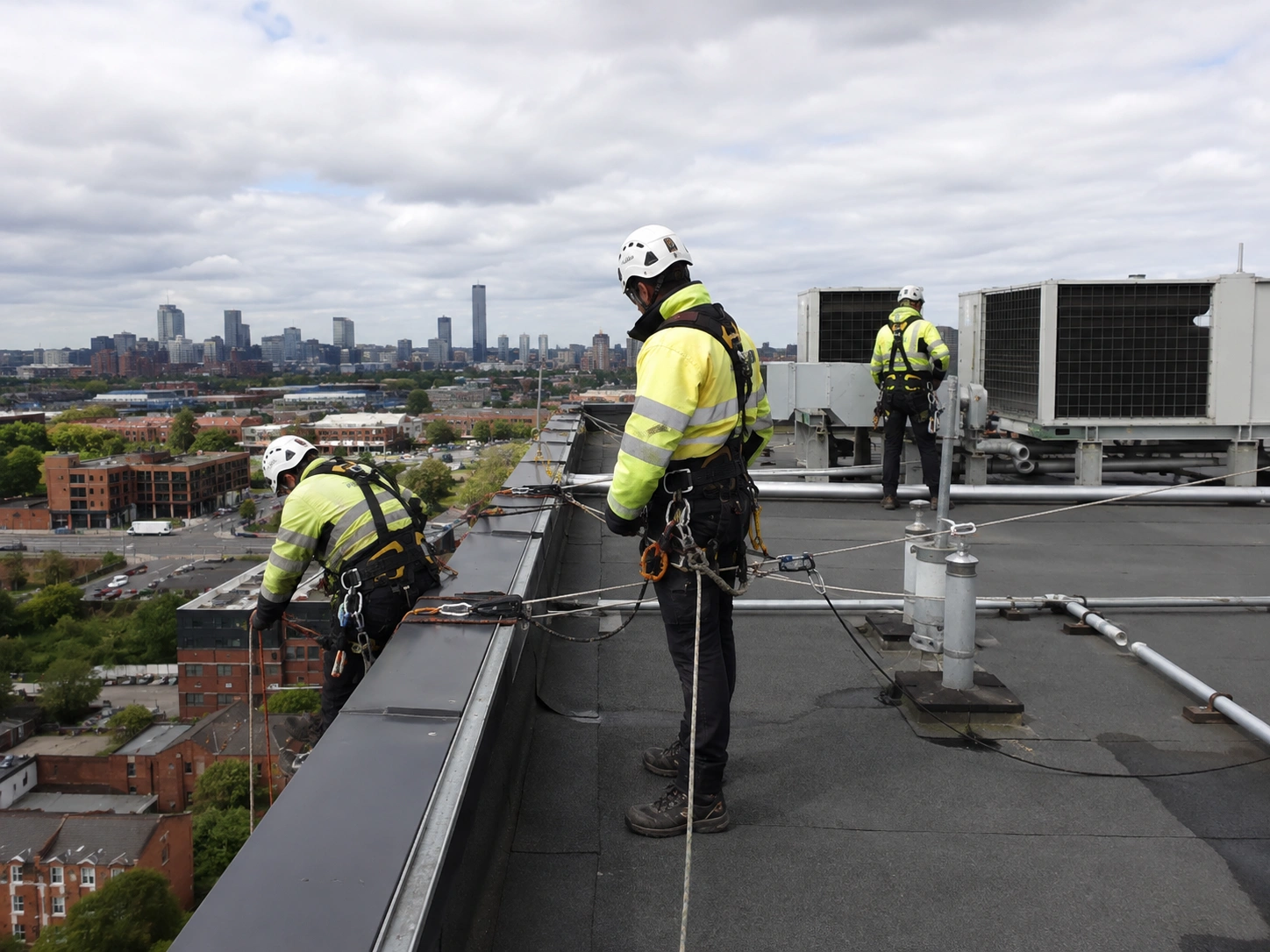 Rope-access technicians working at height on a commercial rooftop with the Greater Manchester skyline behind them